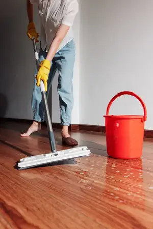 Женщина моет шваброй пол из твёрдой древесины перед окнами, рядом стоит ведро с водой