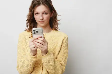 Портрет молодой женщины в желтом вязаном свитере, держащей смартфон обеими руками
