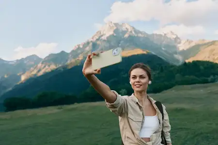 Счастливая женщина делает селфи на смартфон на природе, на заднем плане горный пейзаж, повседневная одежда, яркий дневной свет, расслабленное настроение, летнее путешествие и приключение