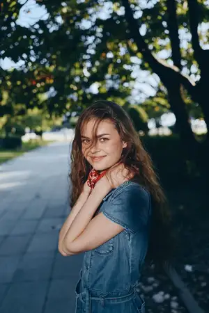 Улыбающаяся женщина в джинсовом платье стоит на затененной парковой дорожке с пышными зелеными деревьями и пробивающимся сквозь листву солнечным светом, излучая тепло и расслабление в летний день