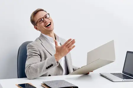 Улыбающийся веселый мужчина в очках и сером пиджаке работает за столом с открытым ноутбуком, документом и смартфоном в современном офисе, выражая радость и успех