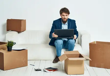 Мужчина сидит на диване перед ноутбуком, работая с коробками вещей, распаковывая новый офис