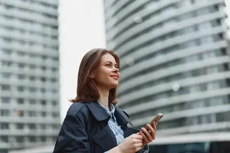 Молодая брюнетка в темном пальто и светло-голубой рубашке держит смартфон и улыбается, стоя на улице у современного изогнутого стеклянного офисного здания под пасмурным небом