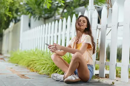 Весёлая молодая женщина сидит на земле, увлечённо пользуясь смартфоном среди пышной зелени и белого штакетника