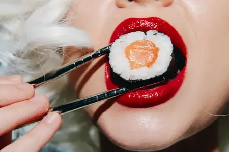 Выразительный крупный план женщины с ярко-красными губами, которая изящно держит суши палочками, воплощая гармонию еды и красоты