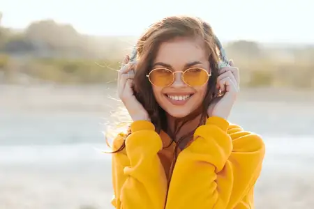 Счастливая женщина в желтом свитере и солнцезащитных очках слушает музыку в наушниках с улыбкой