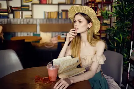 Женщина в шляпе сидит в кафе, перед ней стакан с напитком и книга для отдыха