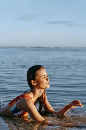 Молодая брюнетка в полосатом купальнике лежит в мелкой морской воде у берега, мягко улыбаясь, пока солнечный свет согревает сцену, а горизонт сияет спокойствием