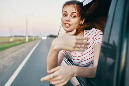 Рыжеволосая женщина в футболке выглядывает из окна автомобиля и жестикулирует руками на летней дороге