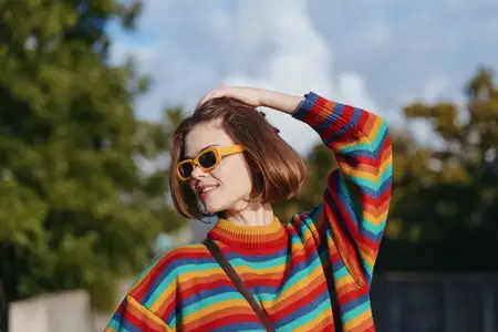 Молодая женщина со стрижкой каре улыбается в красочном полосатом свитере и солнцезащитных очках
