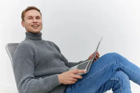 Улыбающийся мужчина расслабляется и работает за ноутбуком, одетый в повседневную серую вязаную водолазку и джинсы, сидит на фоне однотонной белой стены