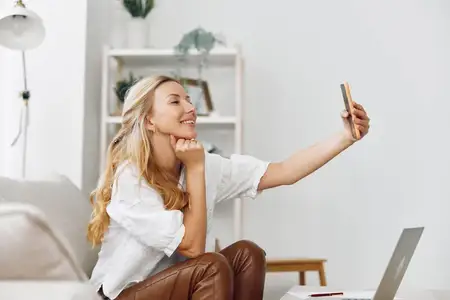 Молодая женщина сидит на уютном диване и делает селфи на смартфон, рядом на столе открыт ноутбук