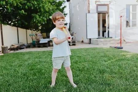 Весёлый мальчик стоит в зелёном саду, держа игрушку, босиком на траве