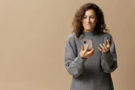 Злая, раздраженная кудрявая красавица в сером повседневном свитере с яростью сжимает телефон, позируя на изолированном бежевом пастельном фоне