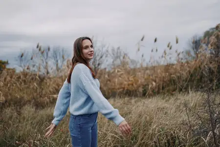 Пасмурное небо и приглушённые тона создают спокойную, достоверную атмосферу, когда она поворачивается с расслабленной позой, передавая реализм, тихую уверенность и непосредственную связь с природой