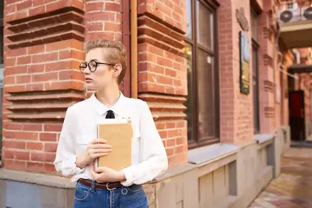 Привлекательная женщина в очках прогуливается по городу с книгой, что символизирует образование