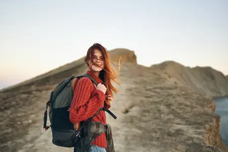 Женщина-туристка с рюкзаком за спиной поднимается на вершину холма вдоль дороги