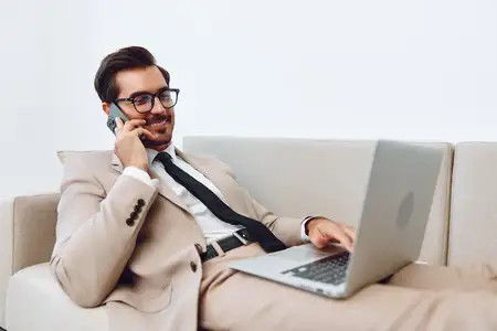Взрослый мужчина в деловом костюме сидит и разговаривает по смартфону, используя ноутбук