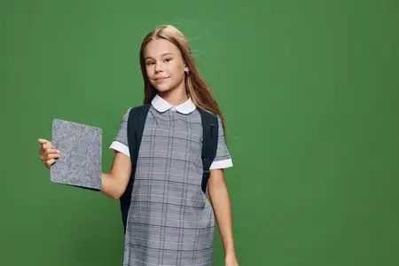 Школьница с рюкзаком в клетчатом платье держит планшет для образования, технологий и обучения, стоя на зеленом студийном фоне с непринужденным выражением и позой