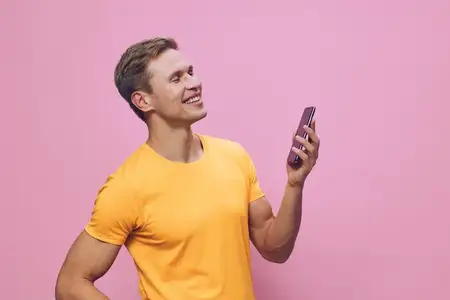 Улыбающийся мужчина держит смартфон в оранжевой футболке, смотрит в сторону с позитивной эмоцией, изолирован на розовом фоне