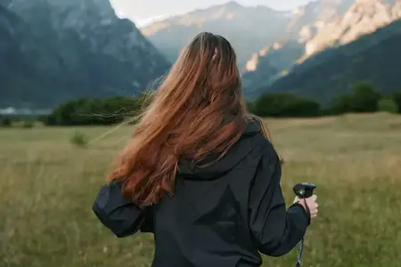 Сцена запечатлела одинокого искателя приключений в походной одежде, держащего треккинговую палку и стоящего на продуваемом ветром лугу, готового исследовать обширные долины и альпийские пейзажи