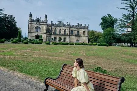 В солнечном парке девушка отдыхает на скамейке возле величественного старинного замка, передавая атмосферу покоя, исследования и вневременной архитектуры