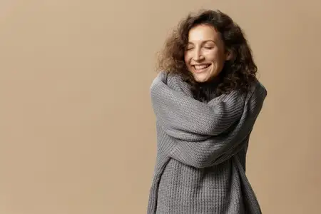 Наслаждающаяся, счастливая, кудрявая, красивая женщина в сером теплом повседневном свитере обнимает себя, закрыв глаза, позирует изолированно на бежевом пастельном фоне