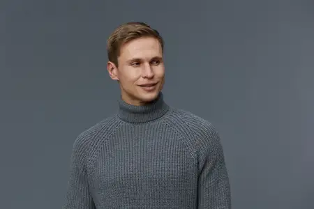 Крупный план портрета улыбающегося молодого мужчины европеоидной внешности в сером свитере с высоким воротником, смотрящего в сторону на темно-сером фоне