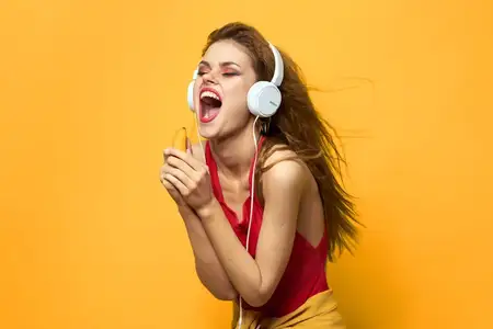 Эмоциональная женщина в белых наушниках слушает музыку, передавая весёлое настроение и стиль жизни на жёлтом фоне
