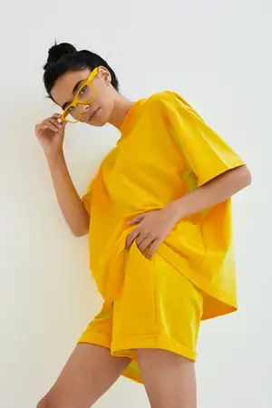 Молодая женщина в жёлтой oversized футболке и шортах уверенно стоит на фоне однотонной белой стены, дополняя образ жёлтыми очками для яркого, современного вида