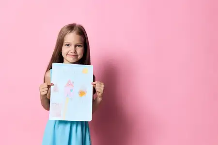 Маленькая девочка в голубом платье улыбается, держа рисунок на розовом фоне, демонстрируя творчество и невинность