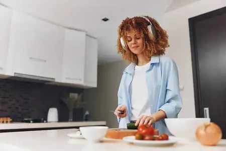 Женщина в наушниках нарезает овощи на разделочной доске на кухне, готовя полезное блюдо