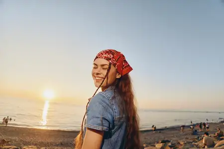 Улыбающаяся женщина, одетая в современный бохо-вестерн деним стритстайл, стоит на пляже во время заката, запечатлевая беззаботный и радостный момент в естественных тонах плёночного цвета