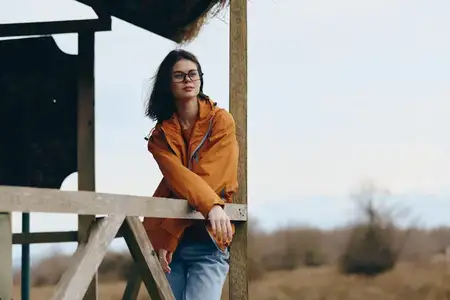 Женщина в повседневном стиле на прогулке: оранжевая куртка и очки, опирается руками на деревянные перила веранды