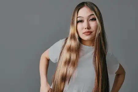 Портрет уверенной молодой женщины с длинными волосами в белой футболке, позирующей с руками на бедрах на фоне однотонного серого фона при студийном освещении
