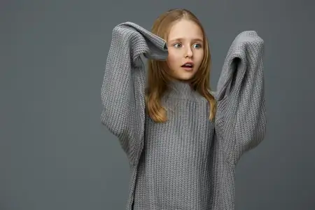 Удивленная юная девушка в oversized сером свитере выражает шок и любопытство на фоне простого серого фона