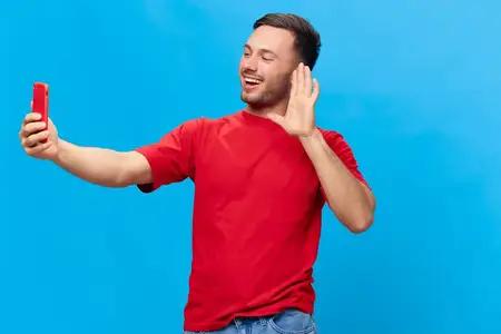 Жизнерадостный, загорелый, красивый мужчина в красной футболке совершает видеозвонок по телефону, приветствуя друзей, и позирует изолированно на синем студийном фоне