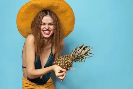 Женщина в купальнике и шляпе с экзотическими фруктами на голубом фоне, излучающая летнее веселье