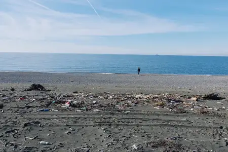 Замусоренный пляж и загрязнённый берег с одиноким прохожим у воды, открытое небо и спокойное море, подчёркивающие экологические проблемы и важность защиты и уборки
