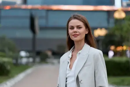 Уверенная женщина в деловой повседневной одежде стоит на открытом воздухе с безмятежным выражением лица, современный городской фон, естественное освещение и мягкая цветовая палитра, передающие…