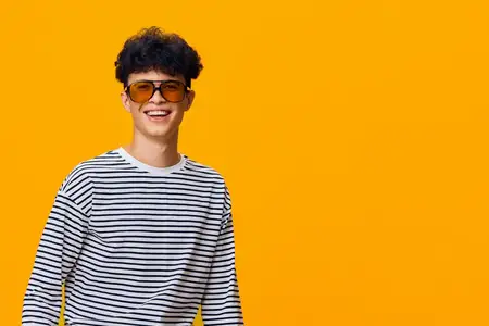 Улыбающийся мужчина в полосатой рубашке и оранжевых солнцезащитных очках демонстрирует юношескую энергию и весёлое настроение на ярко-оранжевом фоне