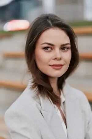 Уверенная женщина в бежевом пиджаке с легкой улыбкой сидит на улице на фоне размытого городского пейзажа, естественное освещение, нейтральные тона