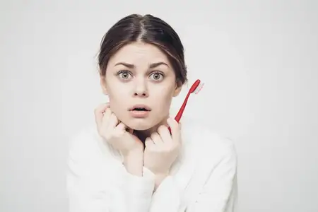 Женщина с зубной щеткой в светлой комнате выполняет утренние процедуры, одетая в халат