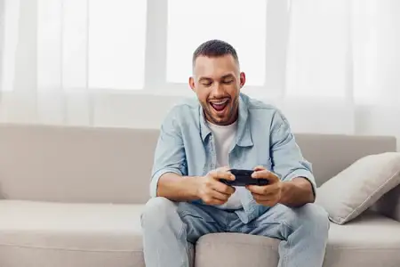 Радостный мужчина занимается цифровым детоксом, играя в игры на своем телефоне в уютной жилой комнате