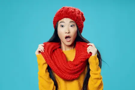Удивлённая азиатская женщина в шарфе и шапке на синем фоне широко открыла рот