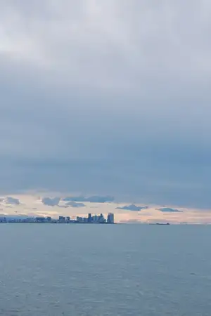 Городской силуэт на фоне облачного неба, далёкий городской пейзаж над широким водным простором с мягким светом и умиротворённым морским настроением