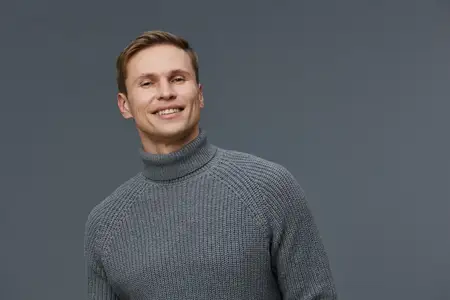 Счастливый мужчина с каштановыми волосами и голубыми глазами, одетый в теплый серый свитер с водолазкой, уверенно улыбается на фоне простого серого фона