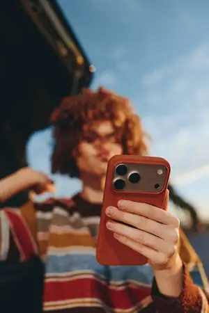 Женщина с вьющимися волосами улыбается, держа смартфон и делая селфи, жизненная сцена на открытом воздухе на городской улице, на заднем плане друзья, дневной свет голубого неба, откровенный портрет