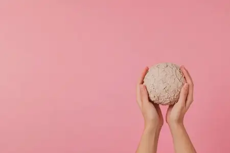 Руки бережно держат текстурированный глиняный шар на пастельно-розовом фоне, подчеркивая темы творчества, искусства и самовыражения
