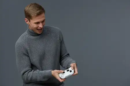 Веселый мужчина в уютном сером свитере играет в видеоигры с игровым контроллером, проявляя волнение и радость на фоне однотонного серого фона, передавая настроение отдыха и развлечения